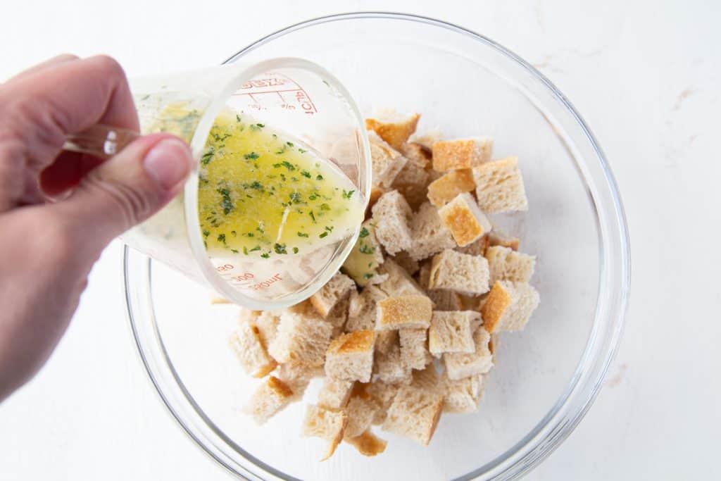 Hand pouring garlic butter over bread cubes in a glass bowl