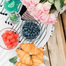 croissants, fresh fruit, and flowers on a picnic blanket.