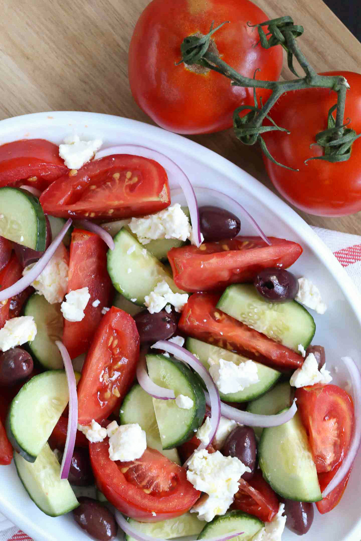 greek salad with feta on a white platter with tomatoes in the background.