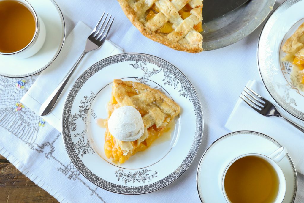 slice of peach pie on a white tablecloth with cups of tea nearby
