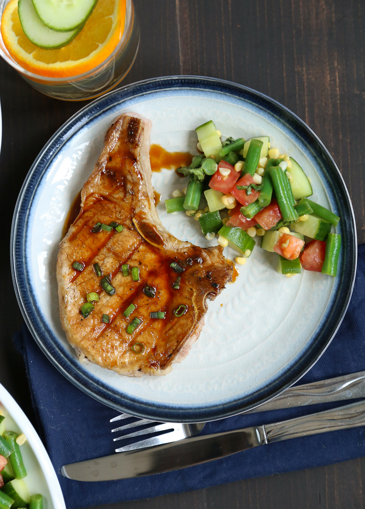 a glazed pork chop with honey and soy, and a summer vegetable salad on a white plate with a blue rim.