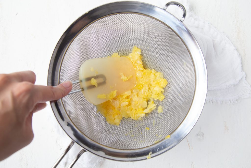 hand with a spatula pressing crushed pineapple in a colander