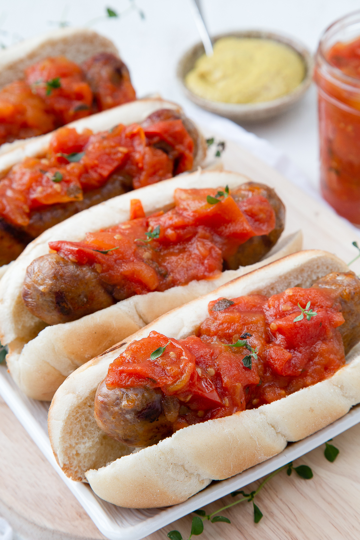 tomato jam on top of bratwursts in buns, on a white serving platter