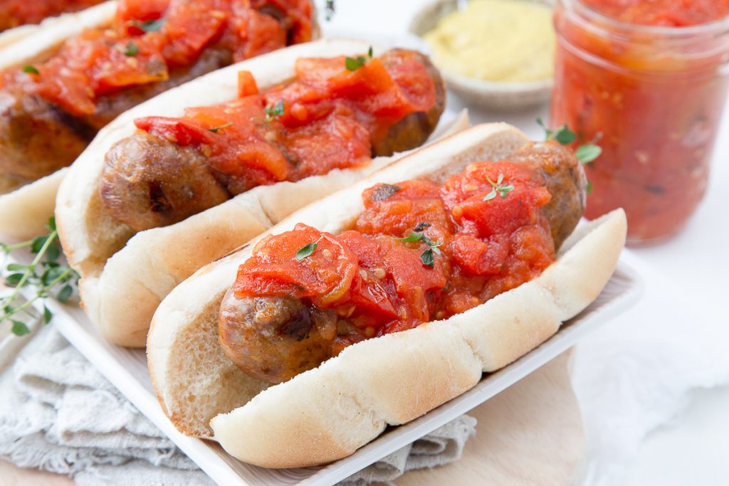 bratwursts on buns topped with tomato jam, sitting on a white platter