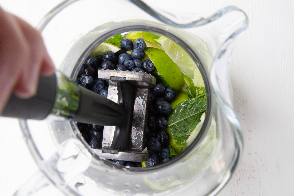 meat mallet smashing blueberries, lime, and mint in a glass pitcher