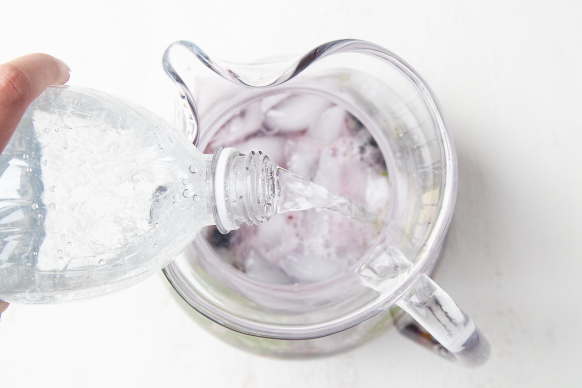 hand pouring club soda into a glass pitcher of mojitos