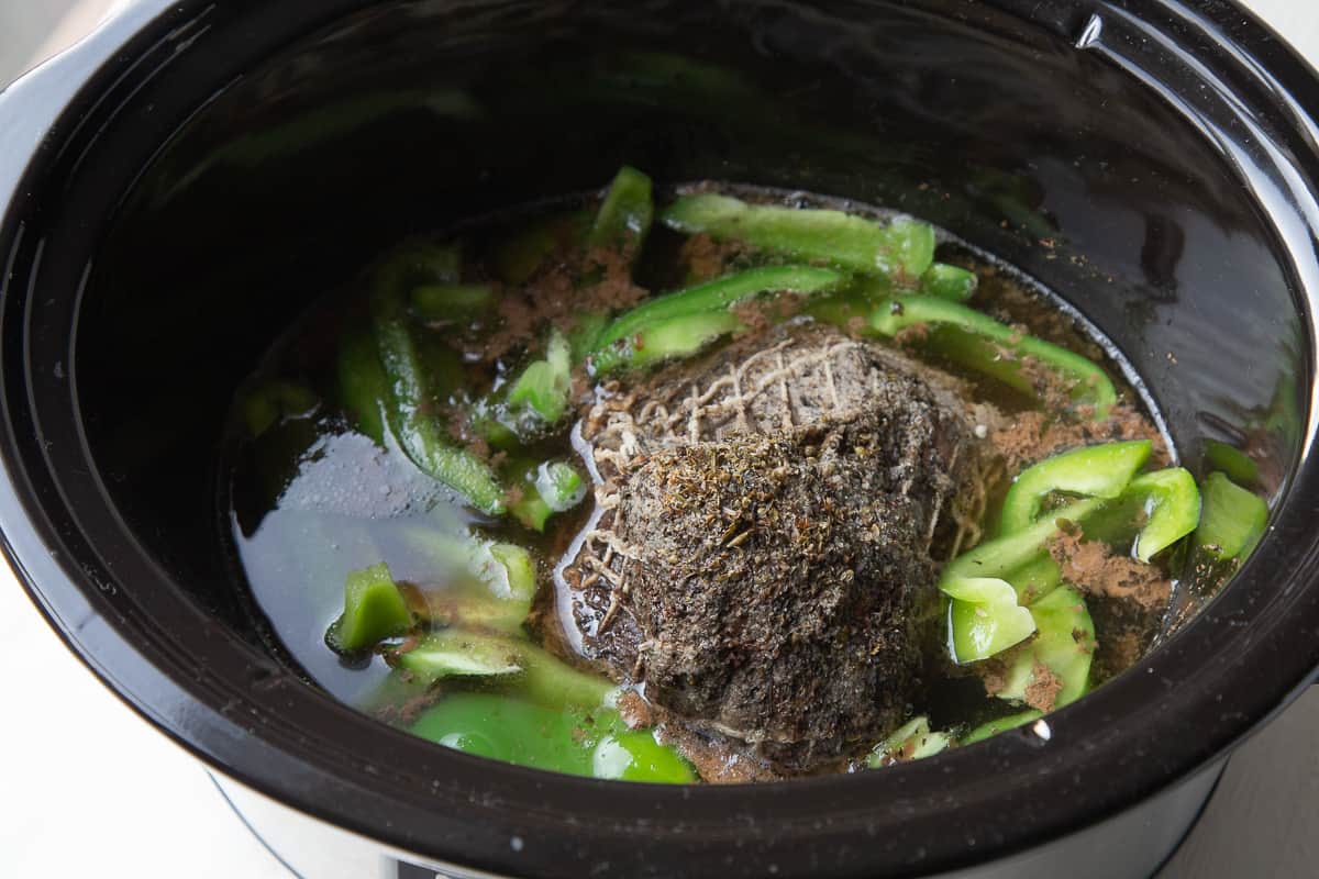 cooked roast and green pepper slices in a slow cooker, sitting in broth.