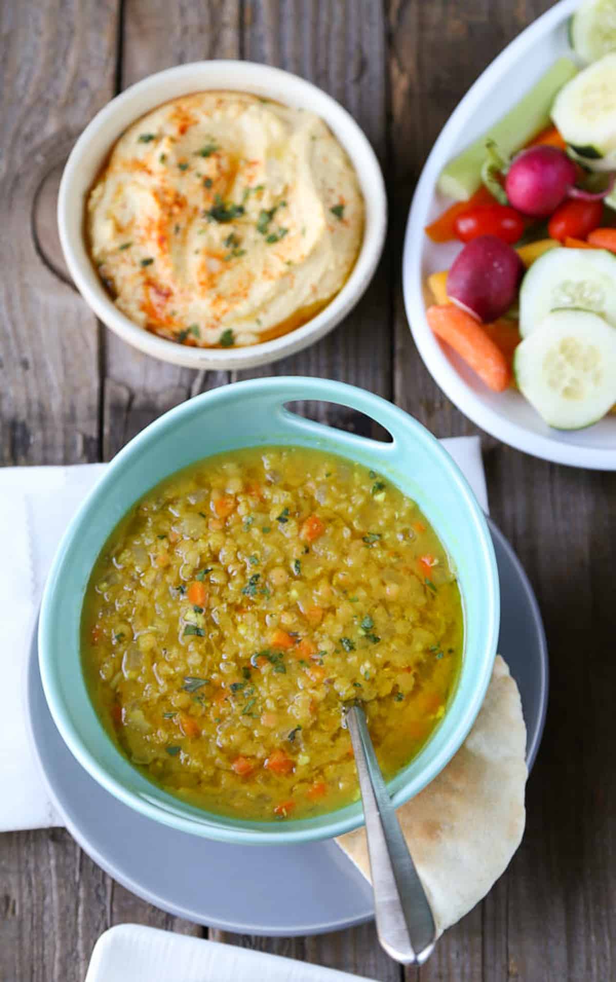 lebanese lemon lentil soup in a turquoise bowl on a wooden table with a side of hummus and raw veggies.