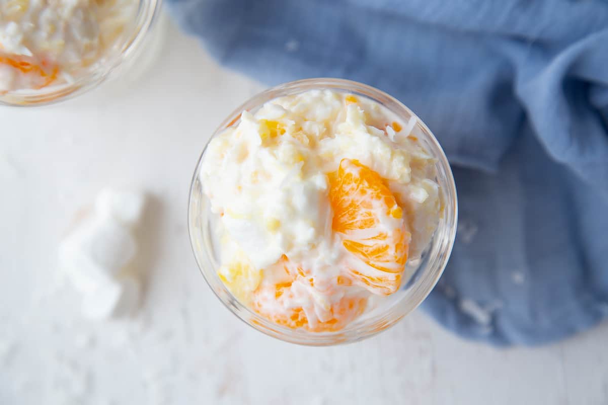 overhead view of a parfait glass filled with ambrosia salad, with a blue tea towel nearby.