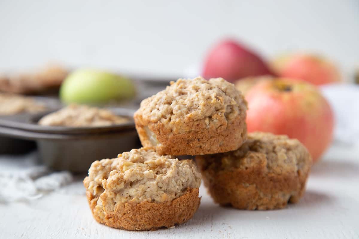 apple oat muffins on a white table with apples and a muffin tin in the background.