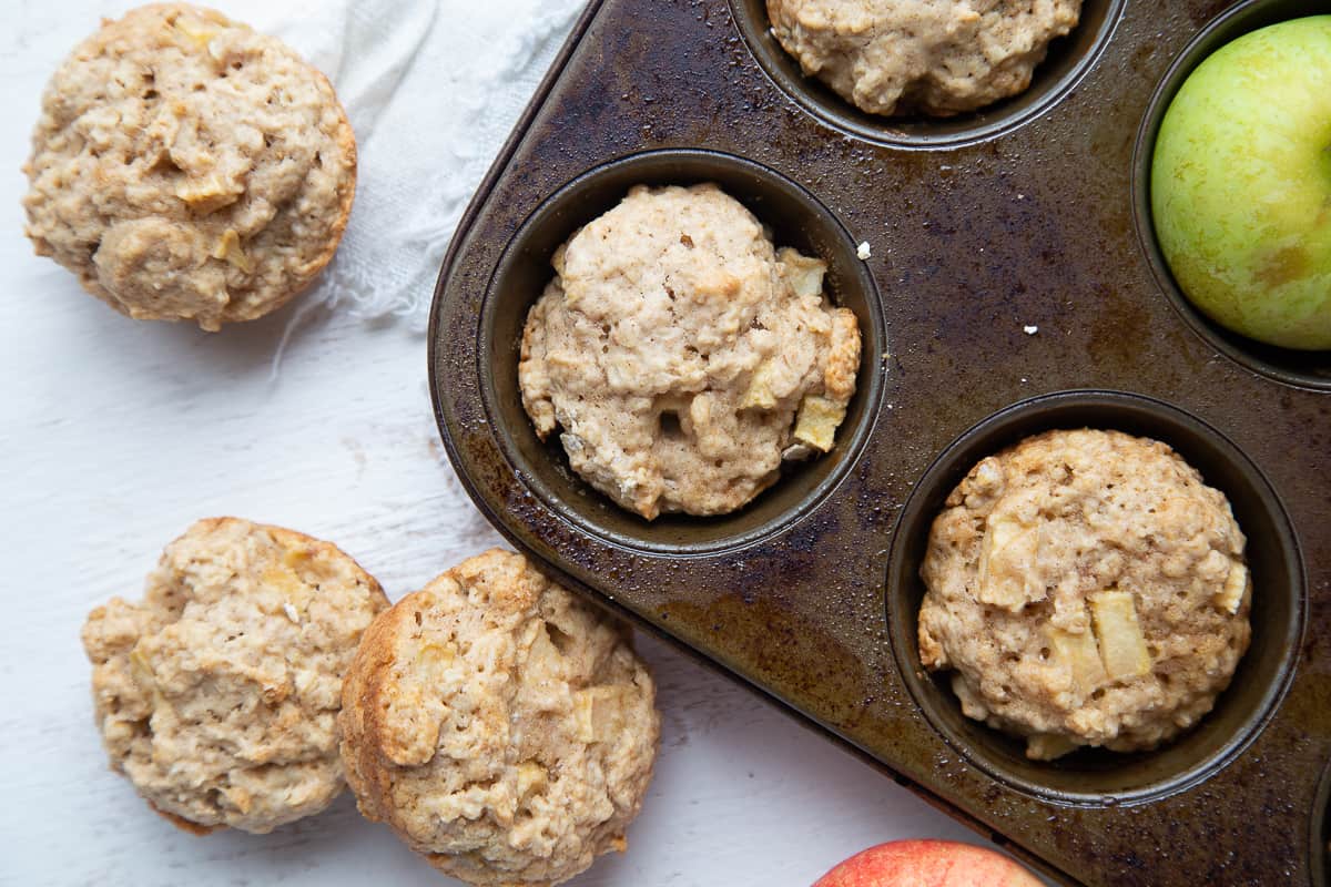 apple muffins in a muffin tin, with more apple muffins on the white table.