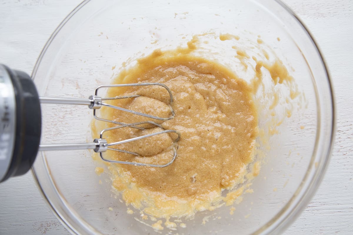 handheld mixer mixing egg, butter, and brown sugar together in a glass bowl.