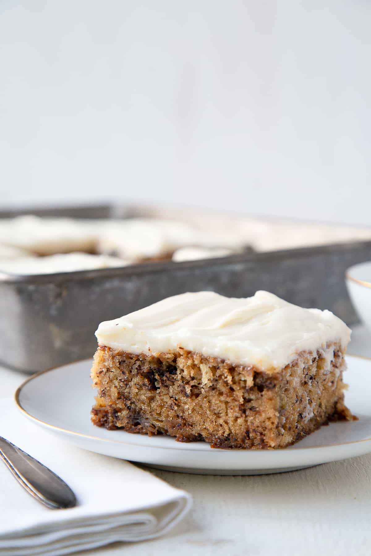 slice of banana cake on a white plate, topped with cream cheese icing with a metal pan of more cake in the background.