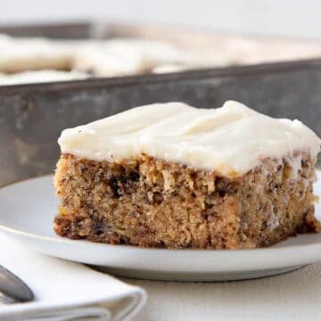 banana cake on a white plate, topped with cream cheese icing with a metal pan of more cake in the background.