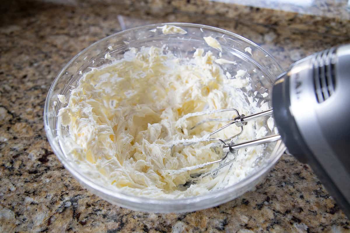 handheld mixer mixing butter and cream cheese in a glass bowl.