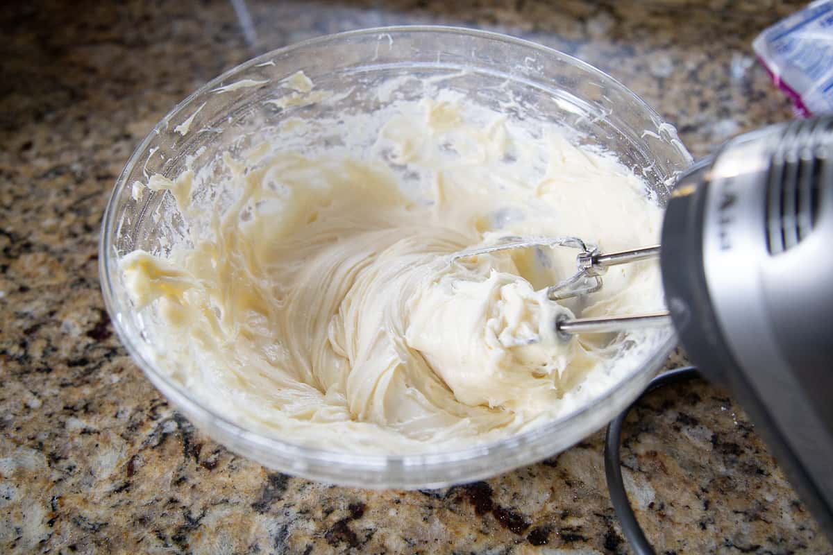 handheld mixer mixing cream cheese frosting in a glass bowl.