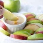 hand lifting an apple slice out of caramel dip, surrounded by apple slices on a white platter.