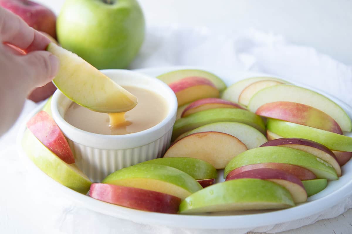 hand lifting an apple slice out of caramel dip, surrounded by apple slices on a white platter.