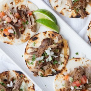 carnitas on top of mini tortillas on a white rectangular platter, garnished with lime slices.