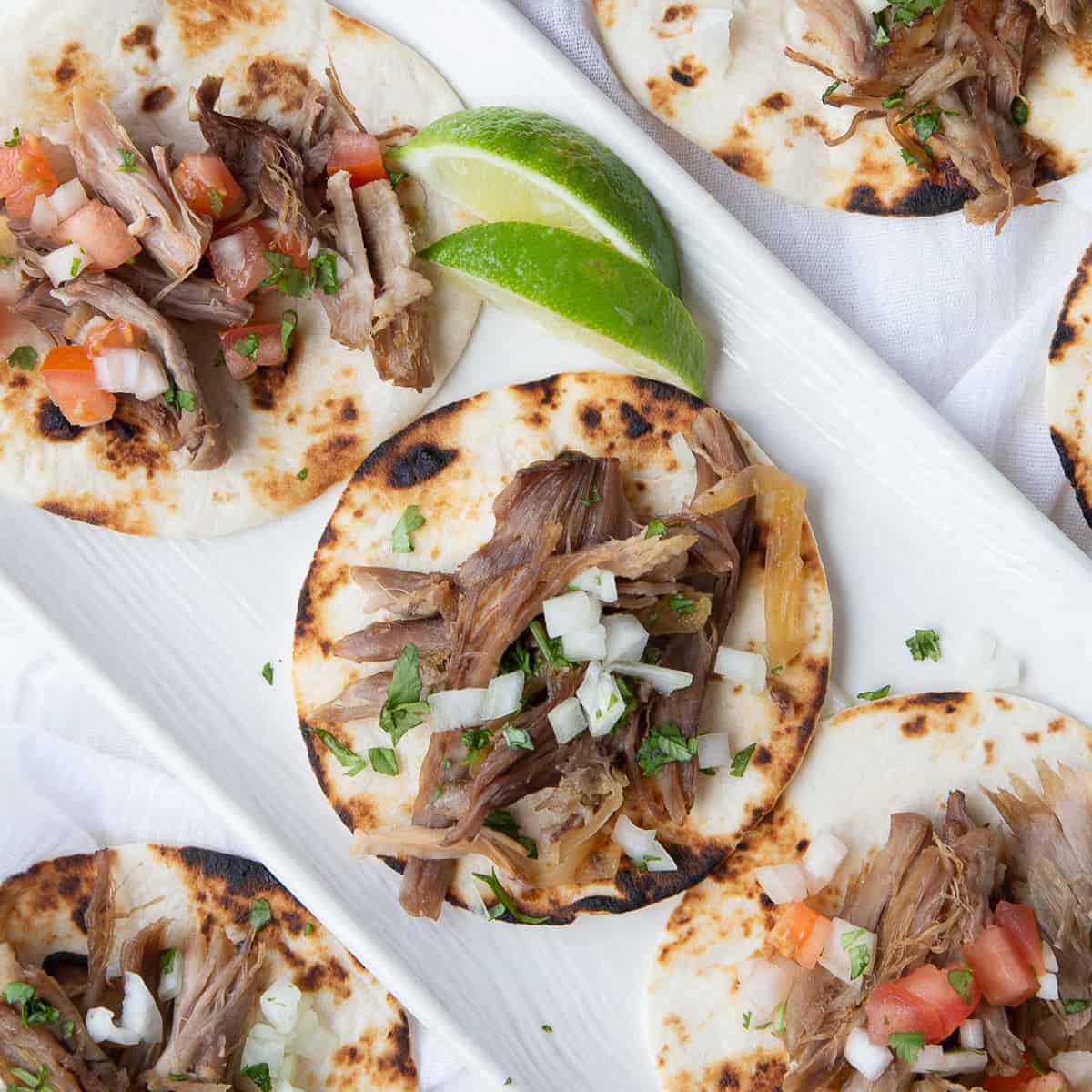 carnitas on top of mini tortillas on a white rectangular platter, garnished with lime slices.
