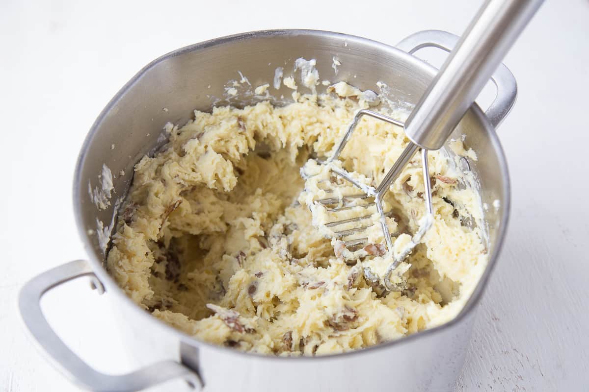 potato masher mashing a large pot of mashed potatoes with the skins on.