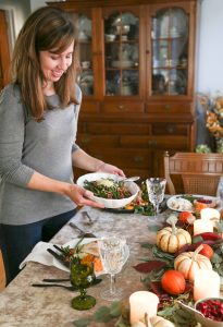 woman bringing a bowl of green beans to a table set for Thanksgiving dinner.