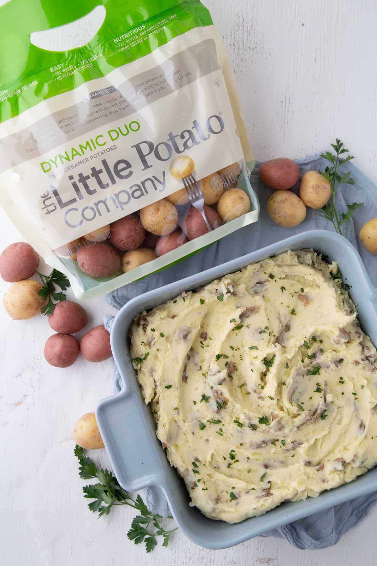 blue square dish of mashed potatoes surrounded by small potatoes and a bag of The Little Potato Company potatoes.