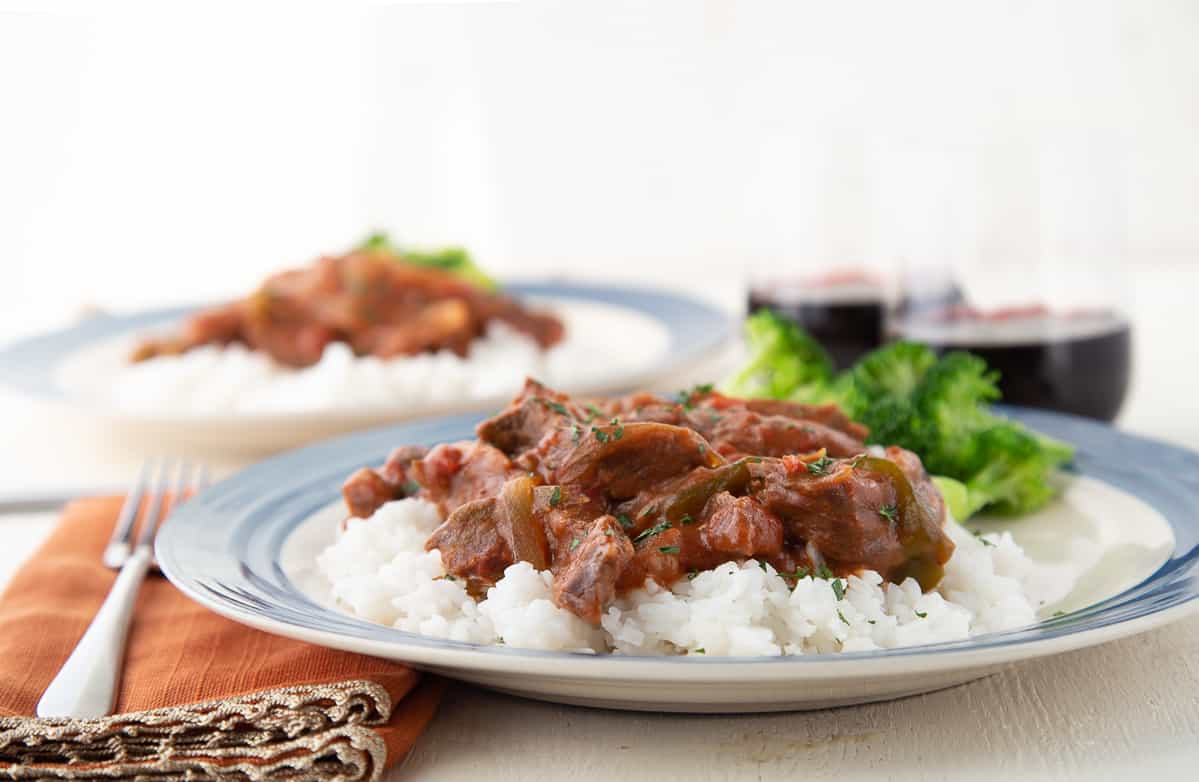 smothered steak on top of white rice on a plate with orange napkins and red wine glasses in the background.