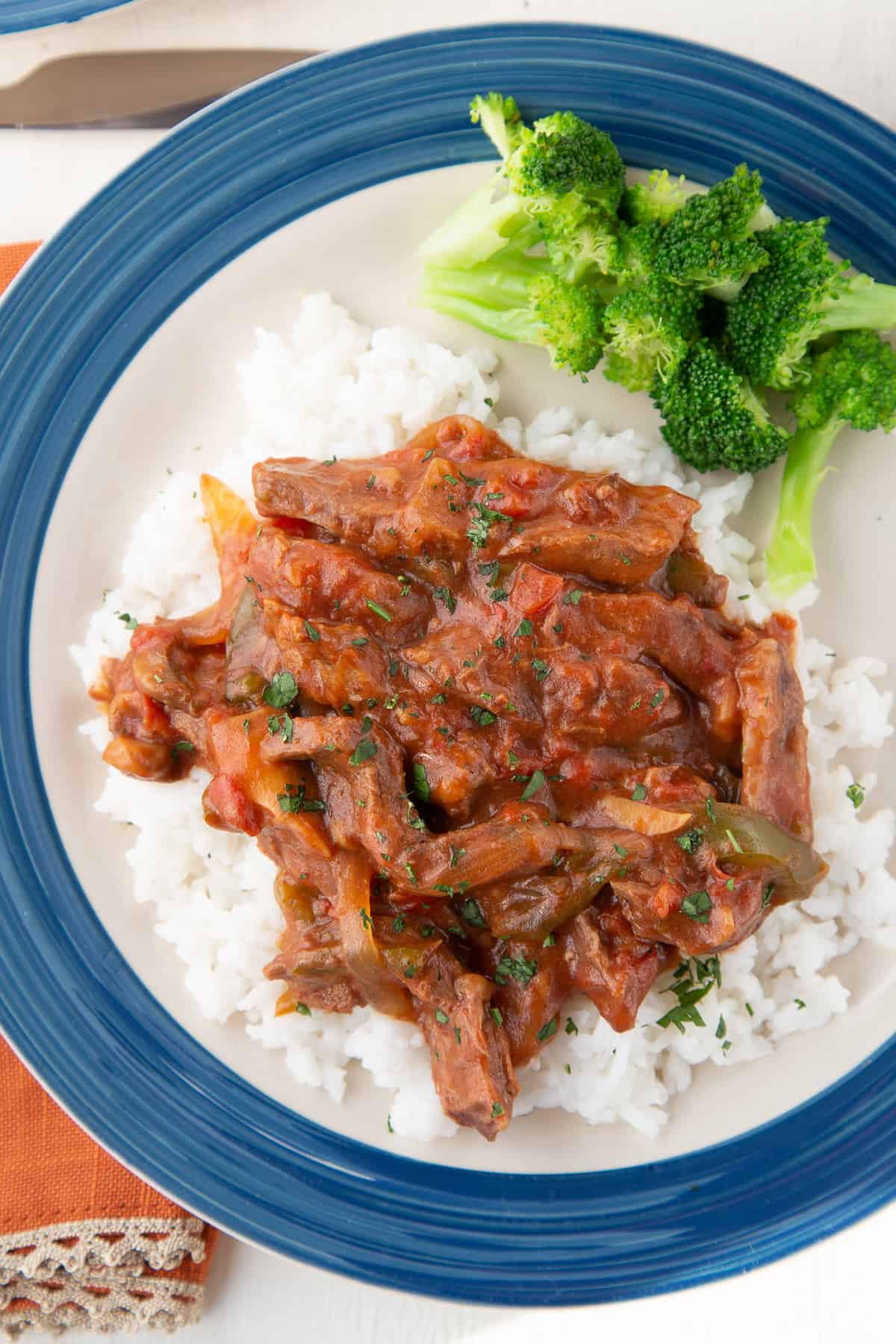 crockpot smothered steak on top of rice with a side of broccoli on a blue rimmed dinner plate.