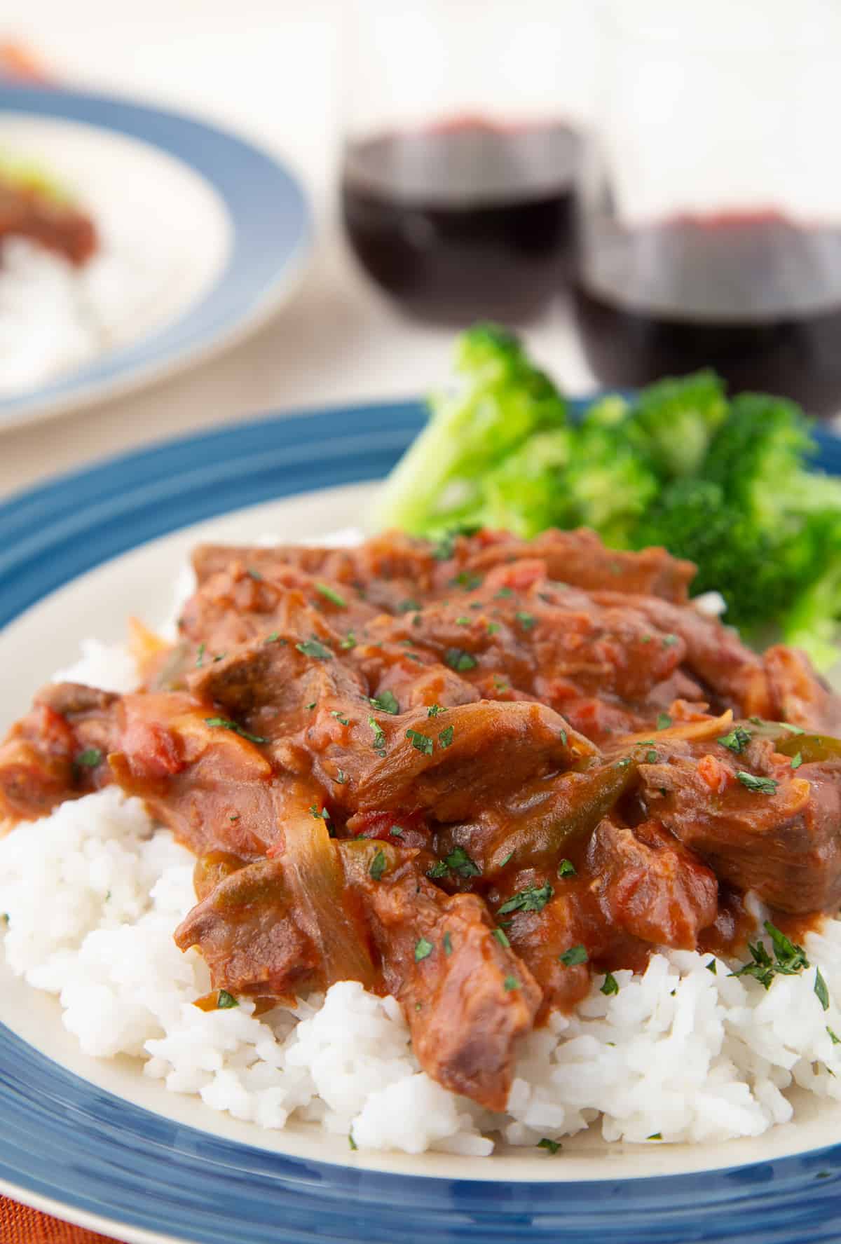 close-up of crockpot smothered steak on top of rice on a blue rimmed plate, with broccoli and stemless red wine glasses in the background.