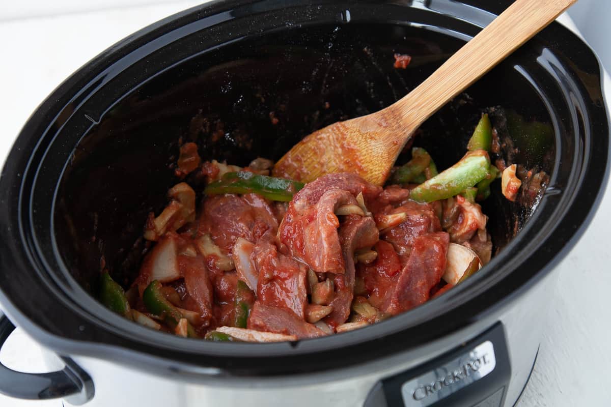 wooden spoon stirring a mixture of tomatoes, veggies, and steak in a slow cooker.