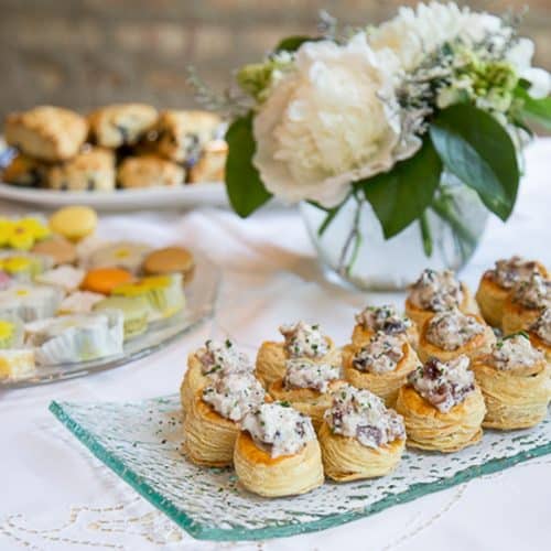 chicken salad in puff pastry cups on a glass serving platter on a buffet table with white flowers.