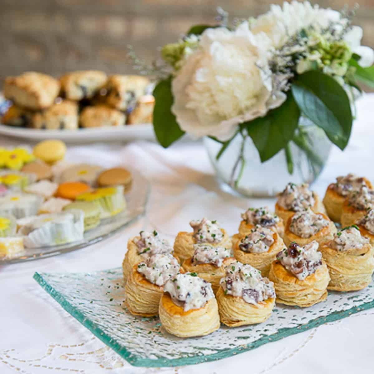 chicken salad in puff pastry cups on a glass serving platter on a buffet table with white flowers.