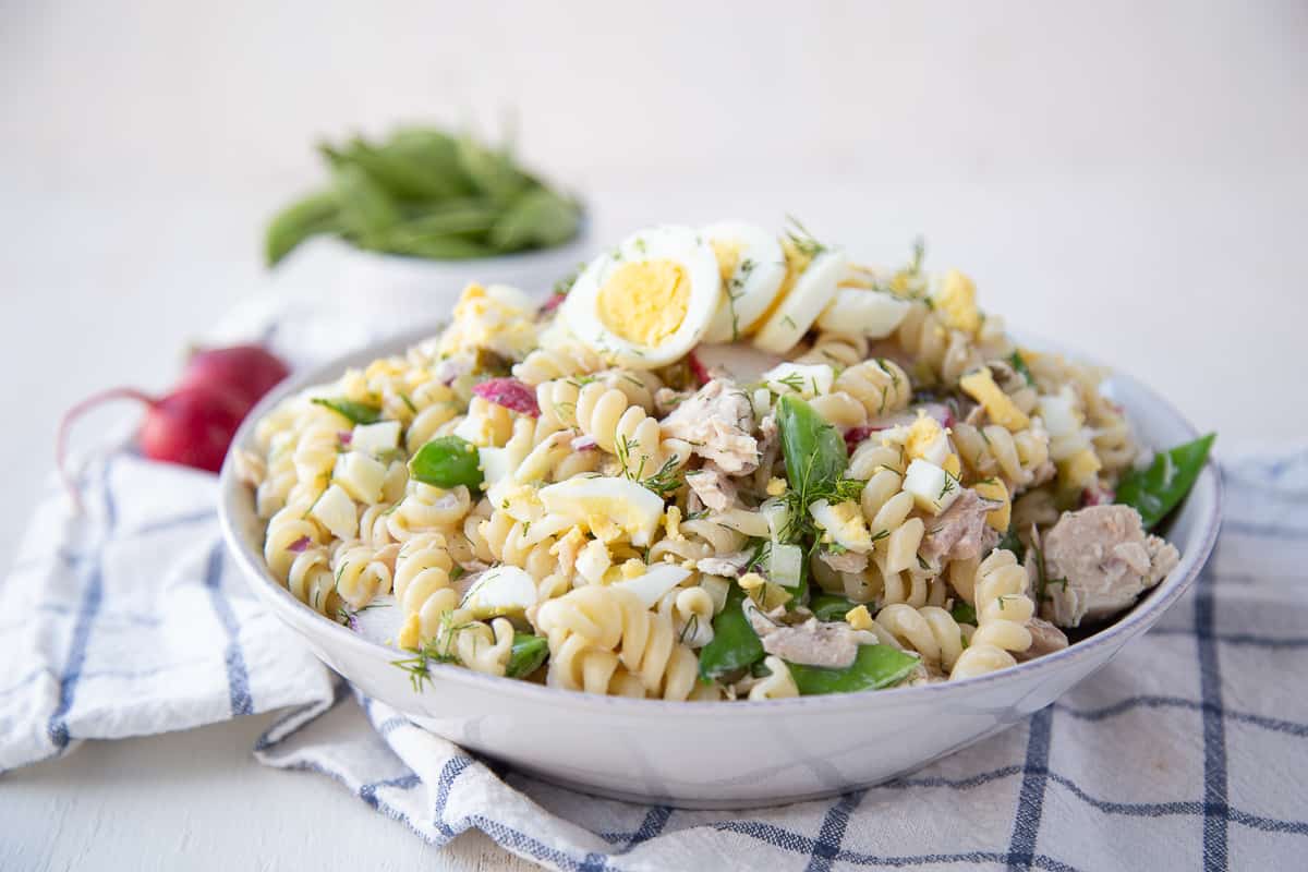 white bowl filled with tuna pasta salad sitting on a white and blue tea towel.