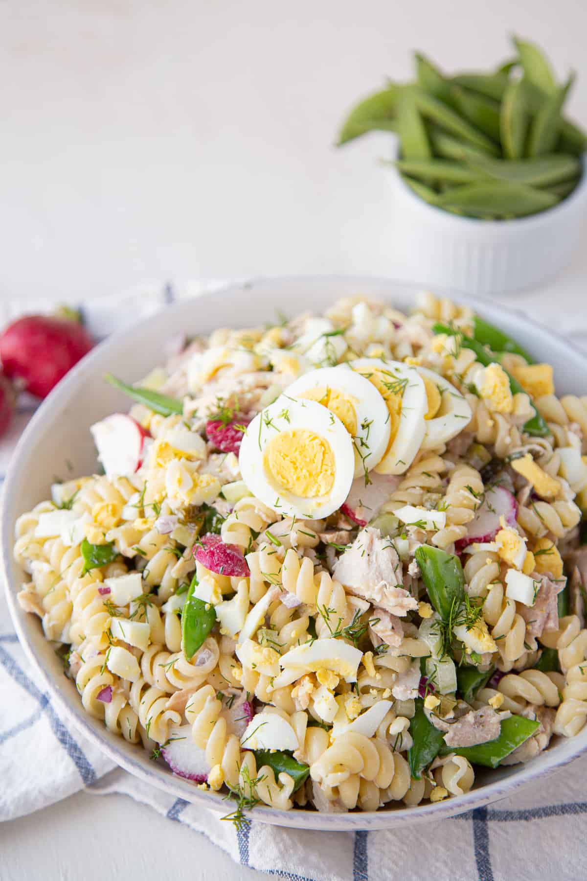 tuna pasta salad in a white bowl with snap peas and radishes on the side.