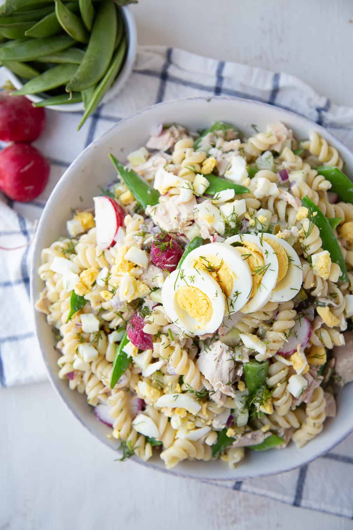 tuna pasta salad topped with hard boiled eggs in a white bowl on a blue and white tea towel.