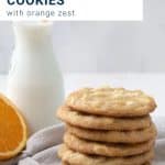 stack of white chocolate chip cookies with a glass of milk and half an orange.