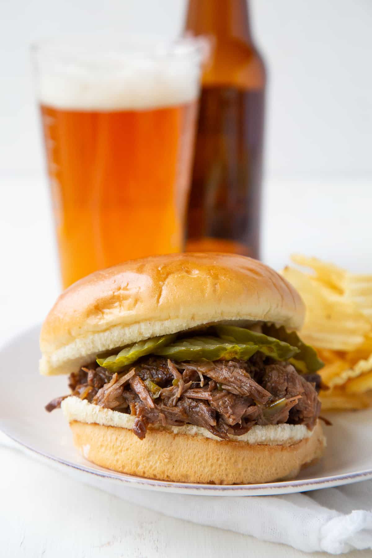 crockpot beef bbq sandwich on a white plate next to a pint glass of beer.