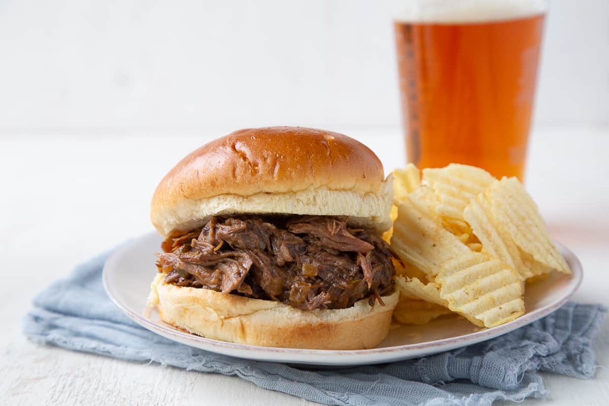 crockpot beef bbq sandwich on a plate with chips and a pint glass of beer.