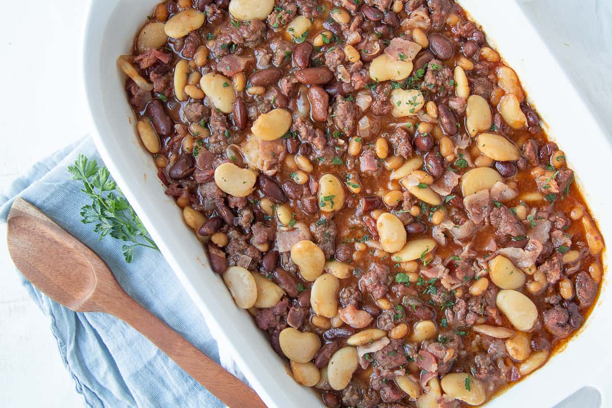 easy baked calico beans in a white casserole dish next to a wooden spoon.