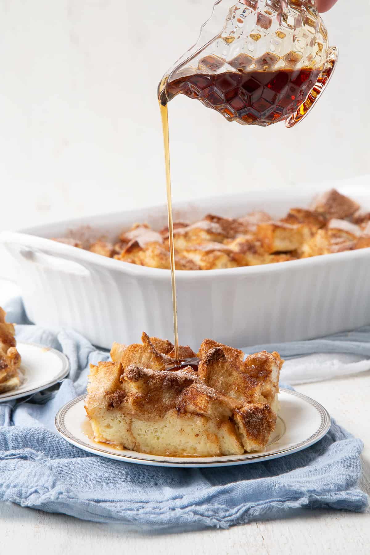 hand pouring a glass jar of maple syrup over a slice of french toast casserole.