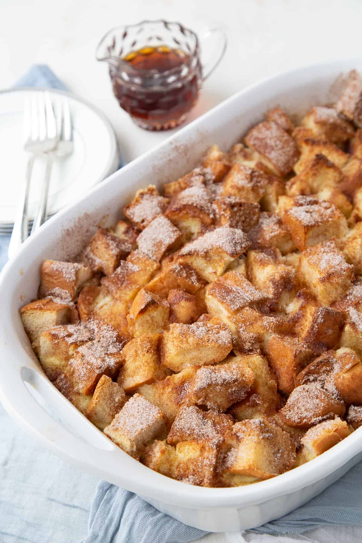 french toast casserole in a white baking dish with a side of maple syrup in a glass mug.