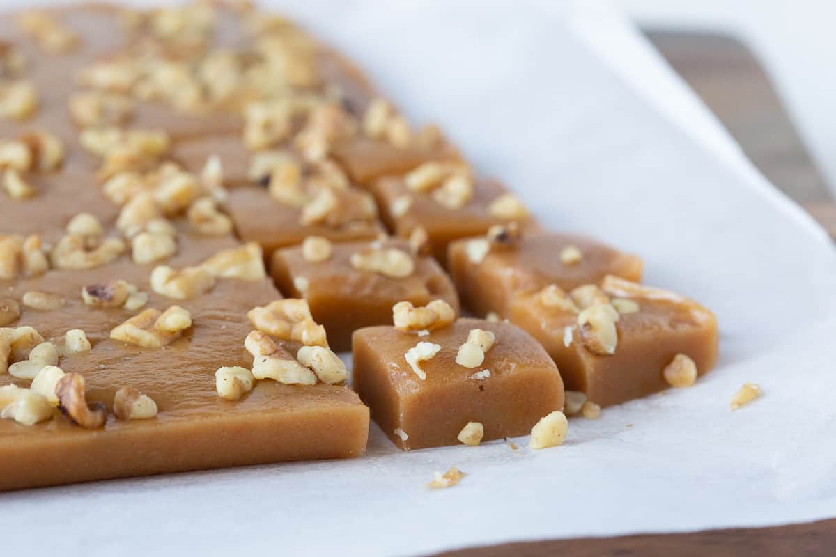 squares of caramels cut from a slab of caramels, topped with walnuts and sitting on parchment paper.