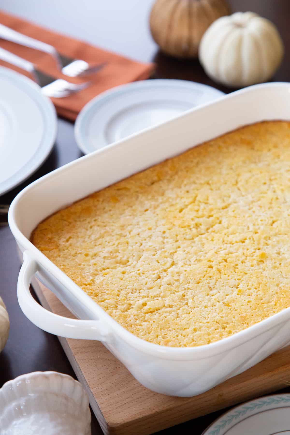 old fashioned corn casserole in a white dish on a table with white pumpkins, white plates, and orange napkins.