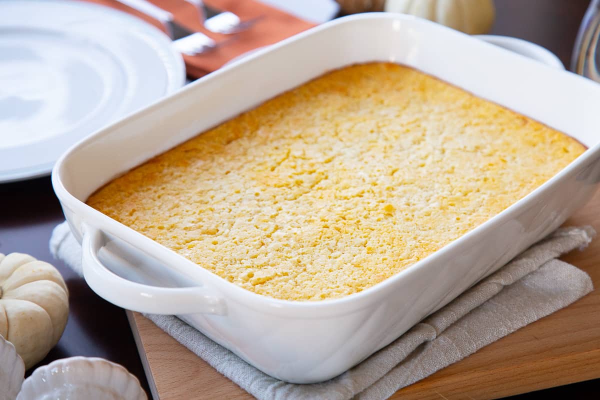 old fashioned corn casserole in a white casserole dish on a table set for dinner.