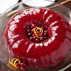 red cranberry jello salad in a ring on a glass cake stand with orange peel and fresh cranberries as garnish.