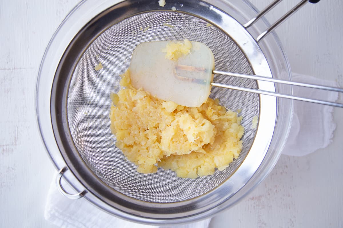 spatula pressing on crushed pineapple in a fine mesh strainer.