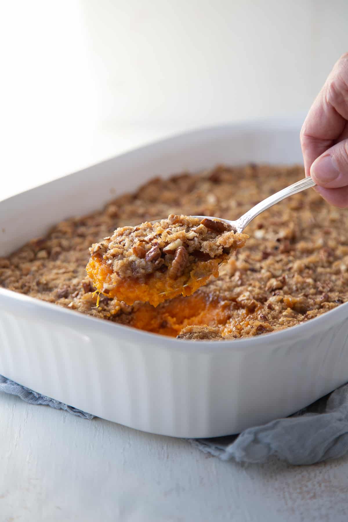 hand lifting spoon of old fashioned sweet potato casserole out of white casserole dish.