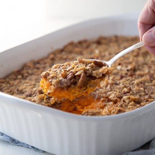 hand with a spoon of sweet potato casserole, lifting it out of a white casserole dish.