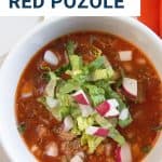 red pozole in a white bowl topped with romaine and chopped radishes.
